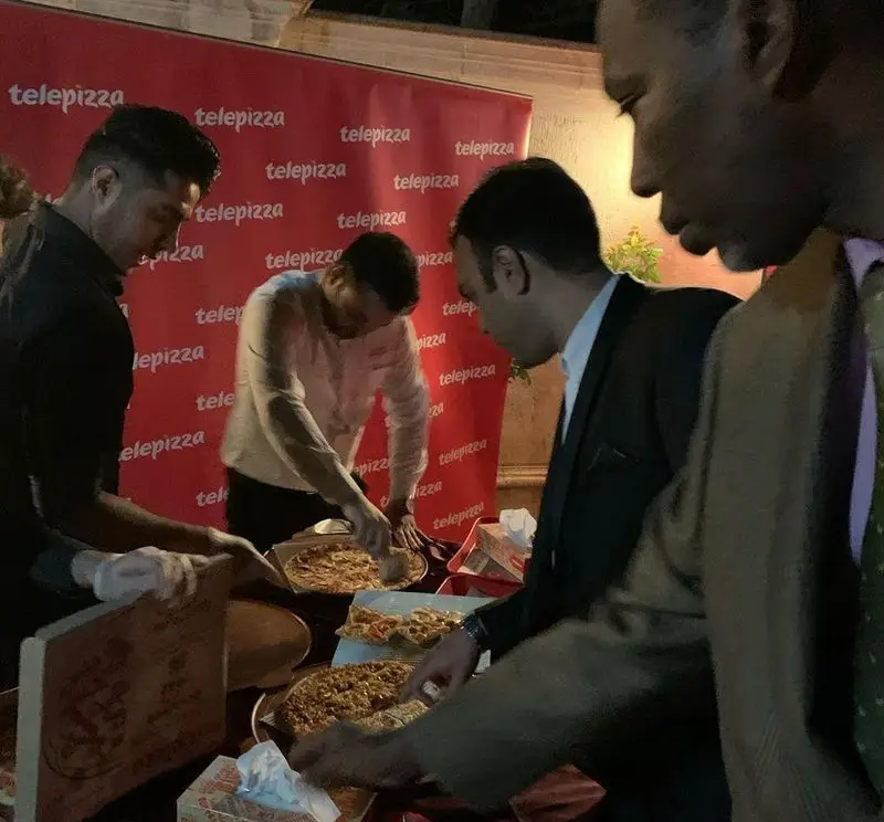 صحنه شلوغ از رویداد تله پیتزا در شعبه پیروزی، افراد در حال سرو و برش پیتزاهای متنوع و تازه در کنار بنر برند.