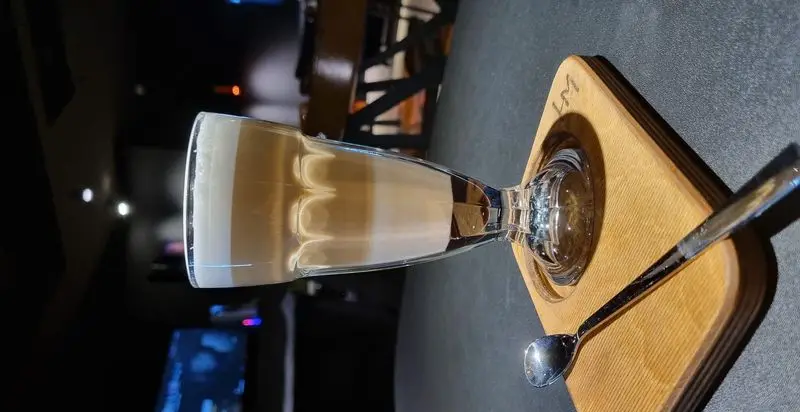 یک نوشیدنی لایه ای خوشرنگ در لیوانی بلند روی سینی چوبی، منتظر شما در محیط آرام کافه گیم استرونات برای لذت از بازی و نوشیدنی.