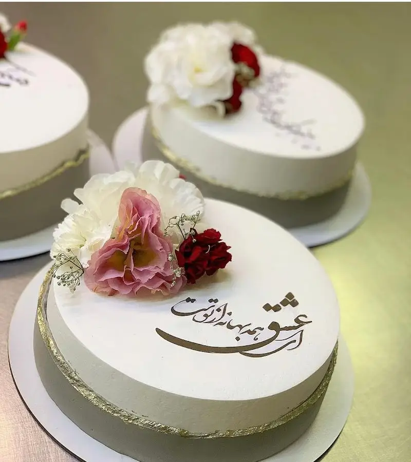 کیک‌های تولد و مناسبتی لوکس با روکش سفید، دیزاین طوسی و طلایی، تزیین گل‌های طبیعی و خطاطی فارسی. شیرینی سرای شکلات تهرانپارس.