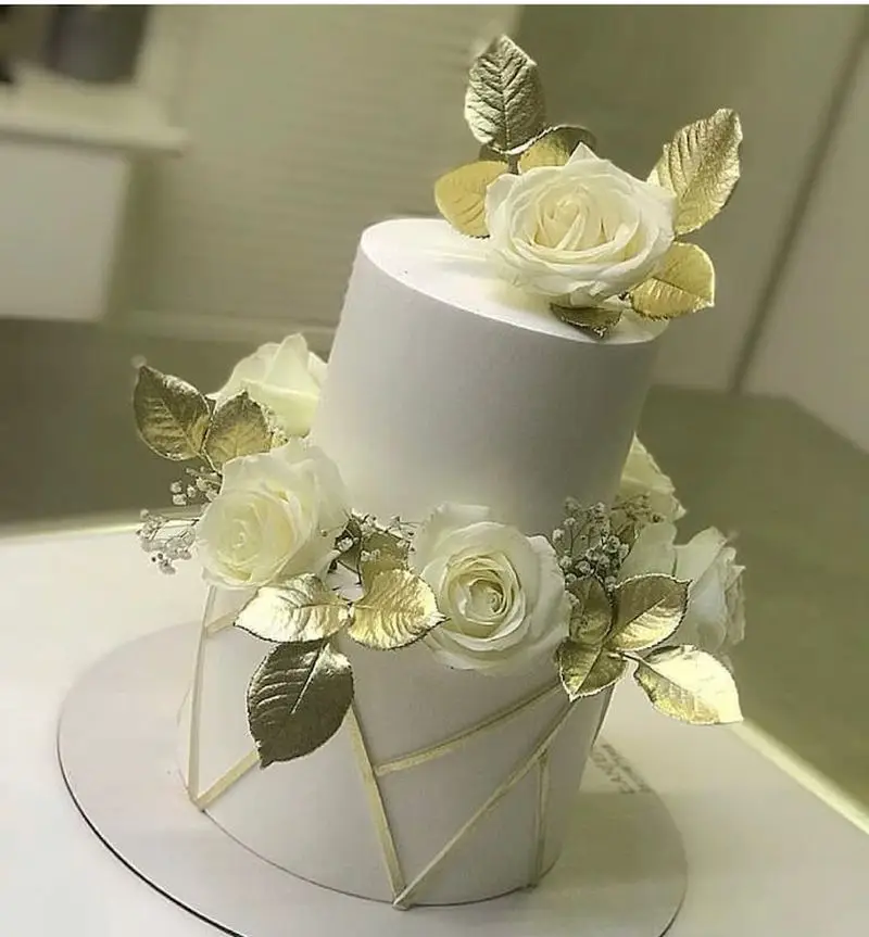 کیک دو طبقه سفید و طلایی لوکس شیرینی سرای شکلات تهرانپارس، مزین به رزهای سفید، برگ طلایی و خطوط هندسی مدرن.