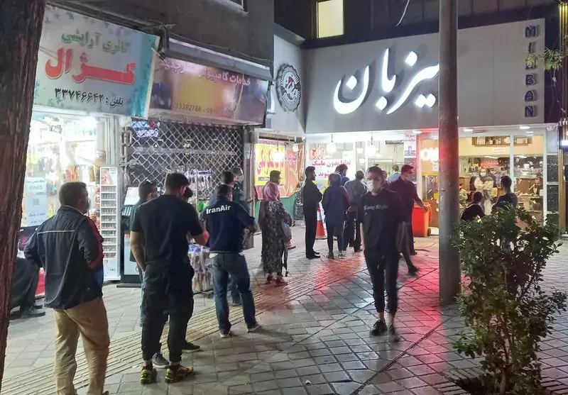 تصویر شبانه از کسب و کار ویتامینه میزبان در خیابان، با حضور مشتریان و رهگذران و تابلوهای روشن مغازه.