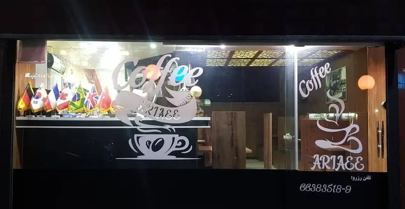 تصویر شب از نمای بیرونی کافه آریایی با نورپردازی، تابلو "Coffee" و لوگوی "ARIAEE" بر روی شیشه ویترین، و نمایش پرچم های بین المللی.
