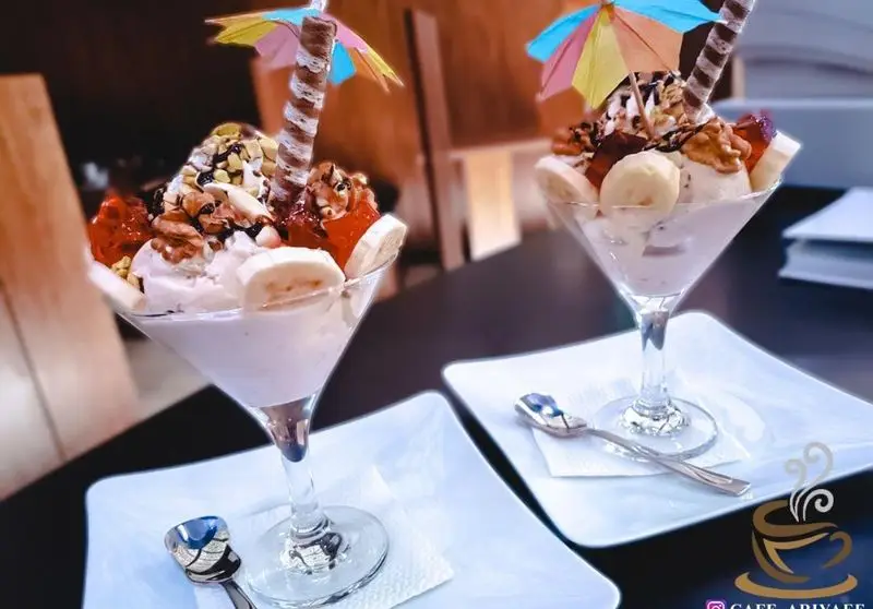 تصویری از دو دسر تابستانی کافه آریایی: بستنی میوه‌ای با موز، توت‌فرنگی، آجیل و سس شکلات در لیوان‌های شیک، آماده سرو.