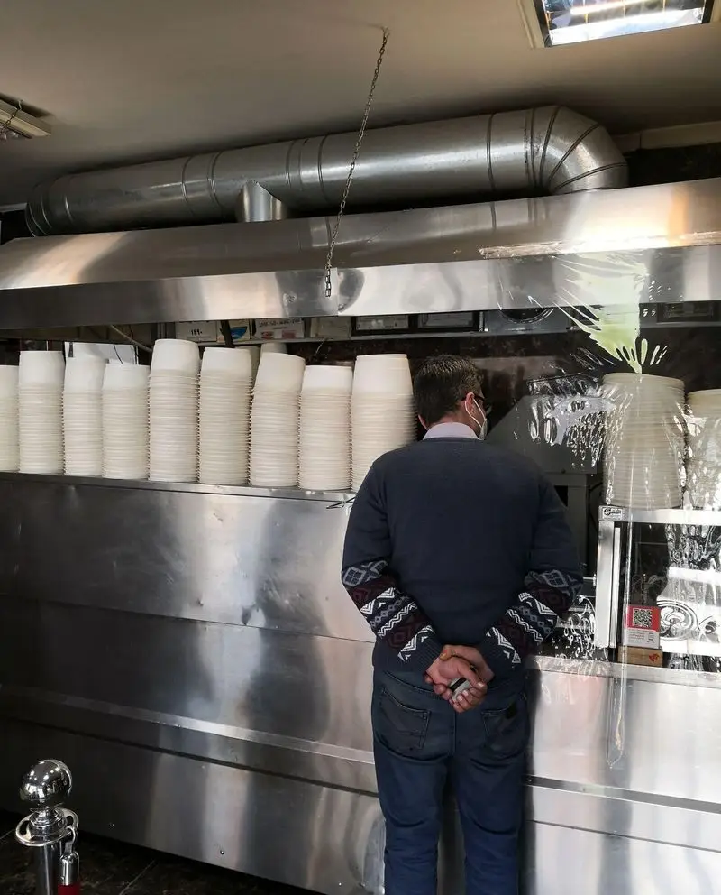 محیط داخلی حلیم فرزین: مردی با ماسک پشت پیشخوان استیل، ظروف یکبار مصرف آماده سرو غذا. بهداشت و نظم در آشپزخانه.