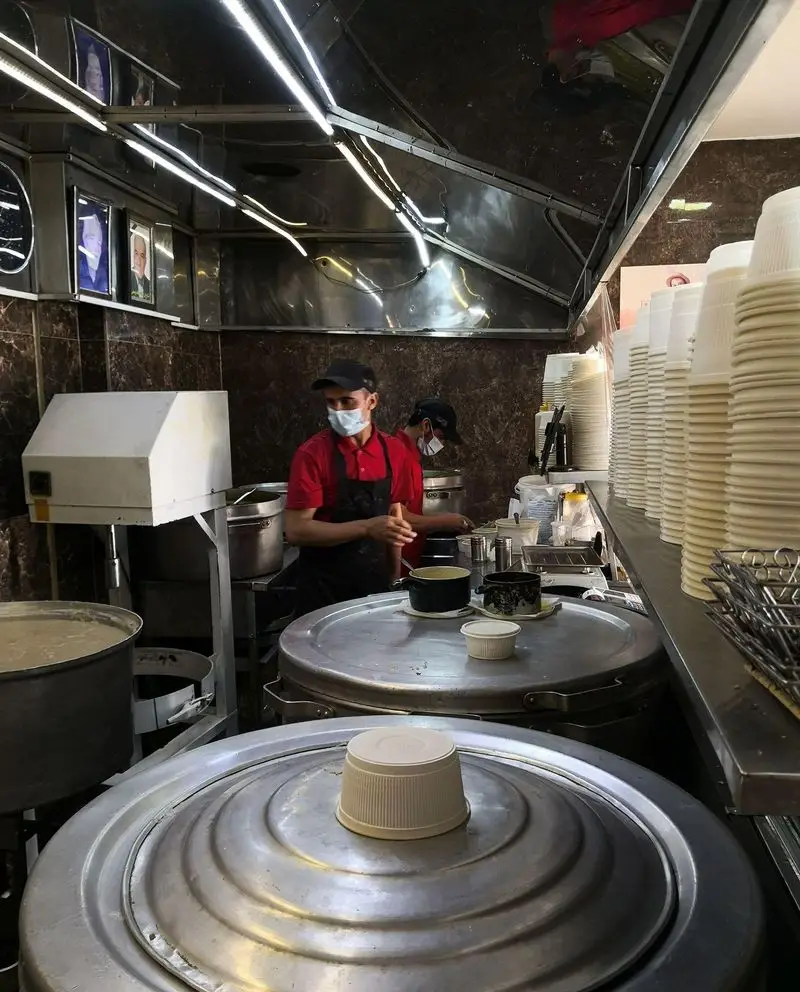 آشپز حلیم فرزین در حال طبخ و آماده‌سازی حلیم سنتی ایرانی تازه و داغ برای سرو به مشتریان در محیطی فعال و بهداشتی.