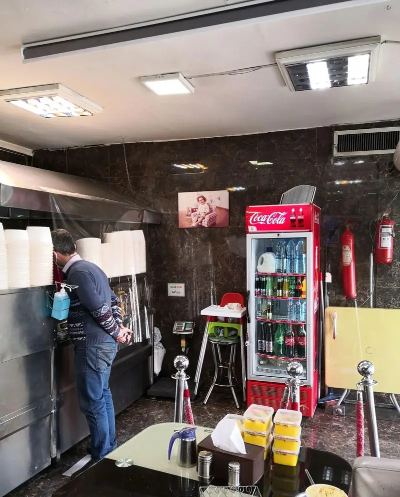فضای داخلی رستوران حلیم فرزین: مشتری در کنار پیشخوان با ظروف غذا، یخچال نوشیدنی، میز پذیرایی و امکانات بهداشتی.