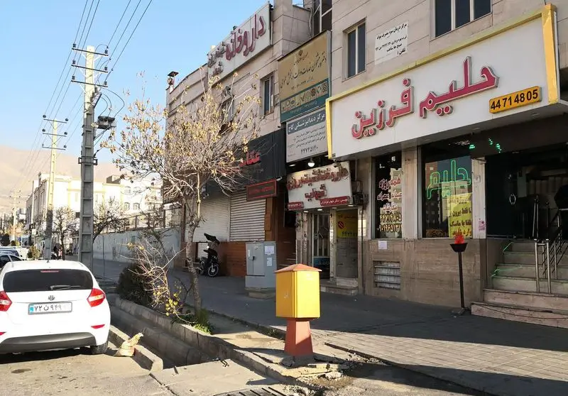 نمای بیرونی و تابلوی درخشان "حلیم فرزین" با شماره تلفن آن در خیابانی پرتردد در تهران، همراه با ماشین و تیرهای برق.