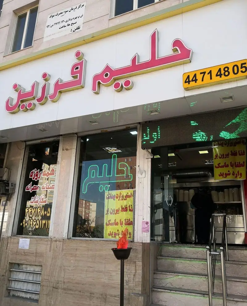 نمای جلوی فروشگاه حلیم فرزین با تابلوی صورتی، شعله نمادین آتش، شماره تلفن و ساعات ارائه حلیم صبحانه و عصرانه