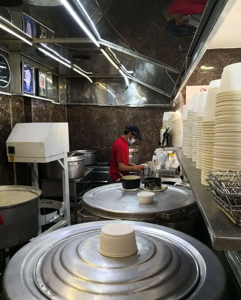 پرسنل حلیم فرزین مشغول پخت و آماده‌سازی غذای سنتی حلیم در آشپزخانه‌ای مدرن با قابلمه‌های صنعتی بزرگ و ظروف سرو آماده.