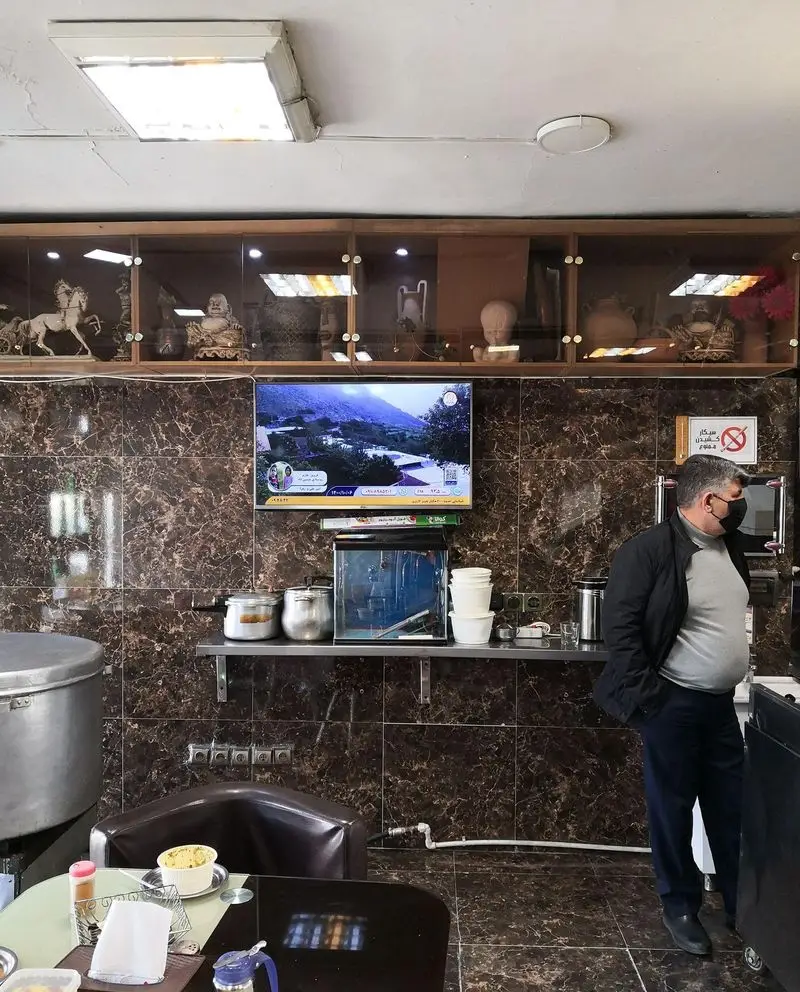 نمای داخلی کسب و کار حلیم فرزین، با دیوارهای طرح مرمر، تلویزیون، ویترین دکوری، قفسه ظروف و مشتری در حال سفارش حلیم.