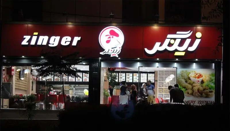 نمای بیرونی شبانه رستوران فست فود زینگر با تابلوهای روشن فارسی و انگلیسی و لوگوی مرغ سوخاری در خیابان.