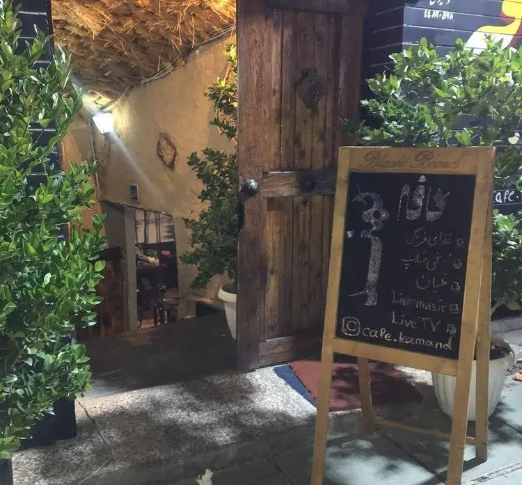 ورودی دنج کافه رستوران کمند با درب چوبی، فضای سبز، و تخته سیاه تبلیغ غذاهای فرنگی، قلیان، موسیقی زنده و تلویزیون.