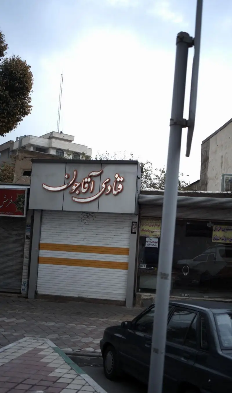 تصویر نمای بیرونی مغازه قنادی آقاجون در یک روز ابری، با کرکره‌های بسته و نوارهای زرد رنگ، یک خودرو در جلوی آن و ساختمان‌های دیگر در پس‌زمینه