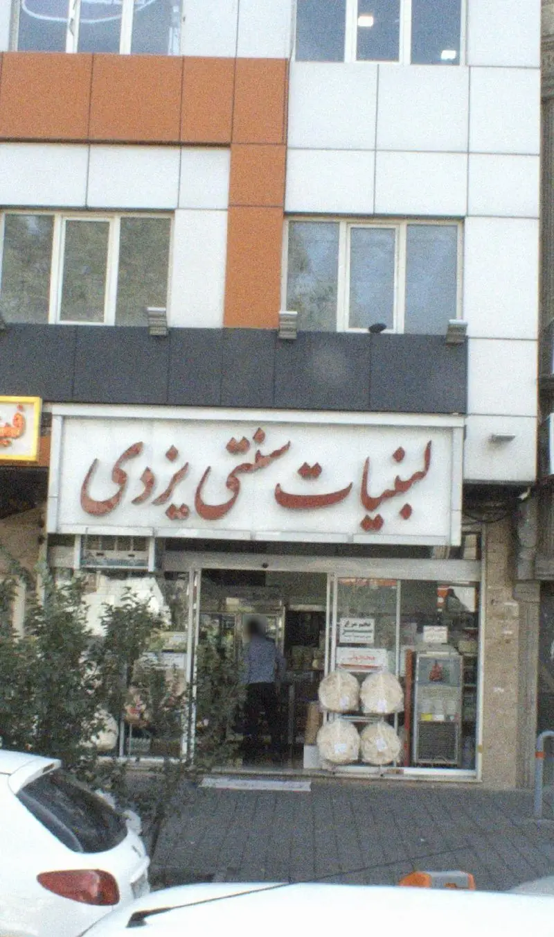 تصویر مغازه لبنیات سنتی یزدی با تابلوی قرمز رنگ و محصولات لبنی سنتی در ورودی، آماده ارائه محصولات تازه.