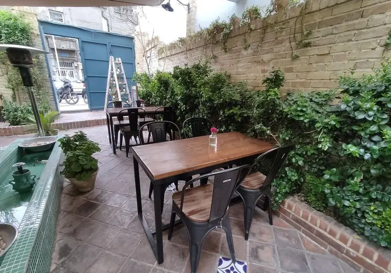 کافه خانه میرا، حیاط دنج و سرسبز با میزهای چوبی شیک، صندلی‌های مدرن، دیوارهای آجری و گیاهان فراوان. محیطی ایده‌آل برای دورهمی.