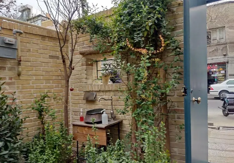 فضای سبز و دلنشین کافه خانه میرا، با دیوار آجری، گیاهان رونده، سینک روستیک و پنجره‌ای دکوراتیو در محیط شهری.