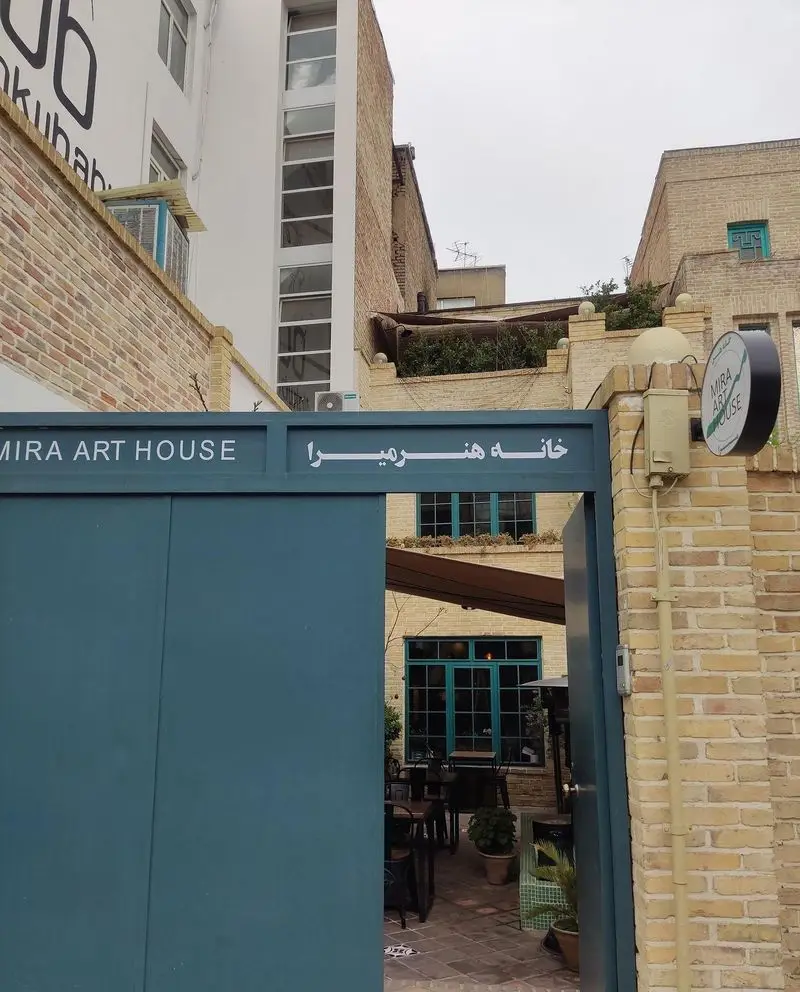 ورودی کافه خانه میرا، با درب سبزآبی، میز و صندلی در فضای باز و پنجره‌های فیروزه‌ای، نشان دهنده یک کافه گالری دنج و هنری.