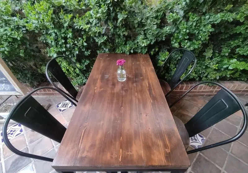 میز چوبی دنج کافه خانه میرا با صندلی های فلزی مشکی در فضای باز و سرسبز، تزئین شده با گلدان کوچک گل.