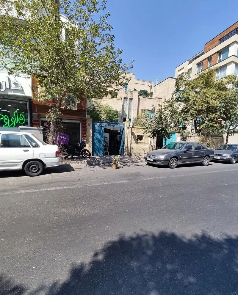 نمای خارجی کافه خانه میرا با درب ورودی آبی رنگ، درختان سرسبز و فضای خیابان در روز آفتابی. محیطی آرام و دعوت کننده برای مهمانان.