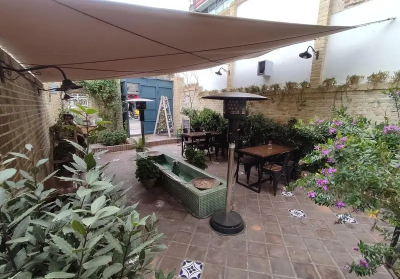 کافه خانه میرا، فضای باز دلنشین و زیبای حیاط با سایبان بزرگ، آبنما، میز و صندلی‌های شیک، گیاهان سبز و بخاری‌های ایستاده.