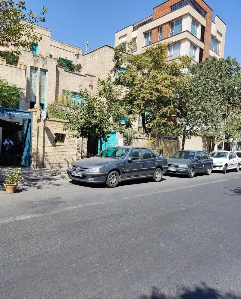 منظره خیابانی کافه خانه میرا در تهران، با ساختمان‌های آجری و نمای مدرن، درختان سبز و خودروهای پارک شده.