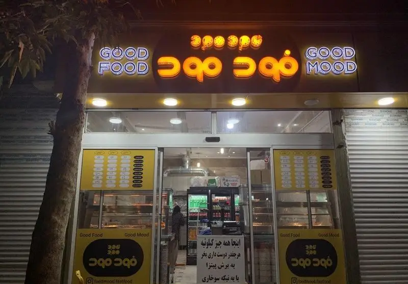 تصویر شبانه فست فود فود مود (Good Food Good Mood) با نمای بیرونی روشن، تابلو نئون و منوی غذاهای متنوع در ورودی.