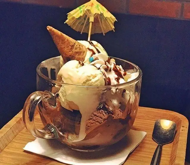 تصویری دلنشین از دسر بستنی لیوانی وانیلی و شکلاتی، با مخروط ویفر، خامه و سس شکلات و چتر رنگی در کافی شاپ گلدون.