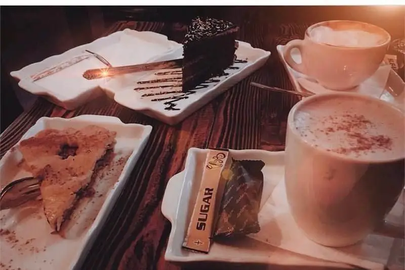 کیک شکلاتی، دسر پای، دو فنجان کاپوچینو و شمع روشن در کافی شاپ گلدون با میز چوبی و فضای دنج برای لحظات شیرین.