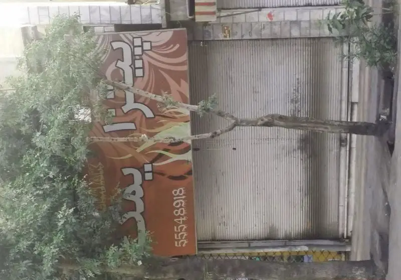 تصویری از نمای بیرونی کسب و کار پیتزا یمک با تابلوی قهوه‌ای رنگ و شماره تلفن 55548918، در حال حاضر با کرکره بسته و درختی در جلو.