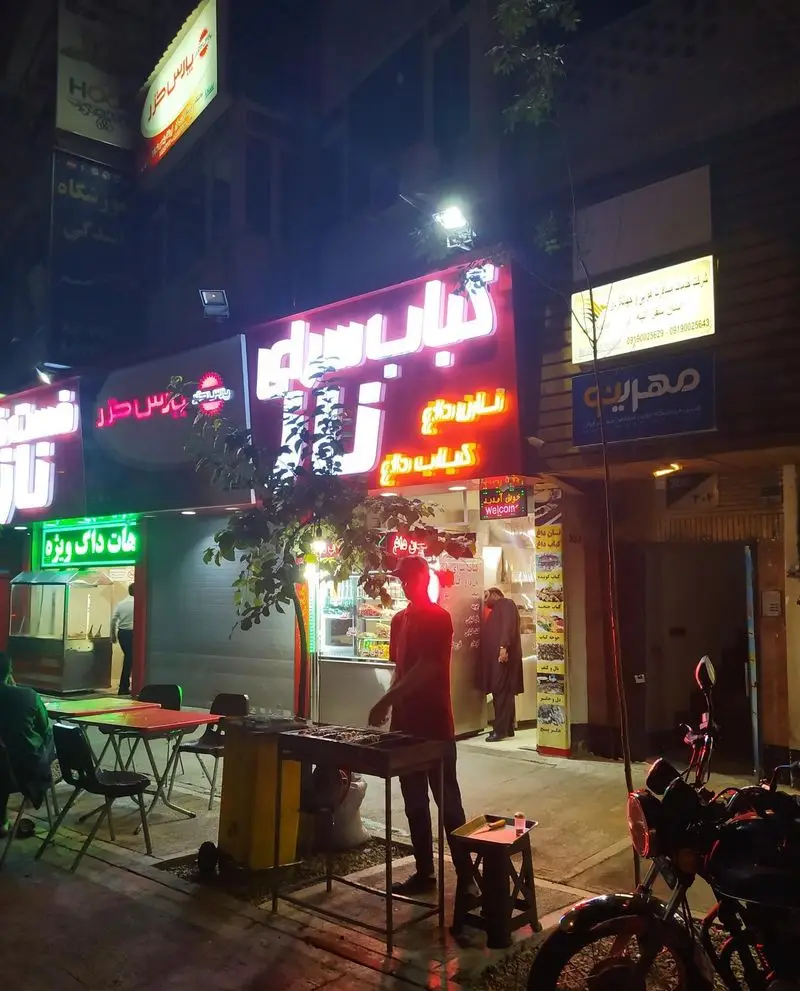نمای شبانه از کباب سرای ناز، با آشپزی در فضای باز و کباب داغ، شامل تابلوی "نان داغ کباب داغ" و مشتریان.