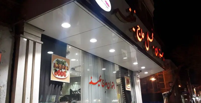 نمای بیرونی روشن رستوران مجلل حاج خلیفه در شب، با تابلوی نئون قرمز و پوستر غذای ایرانی مانند کباب و گوجه.