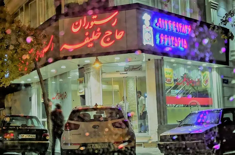 تصویری از نمای بیرونی رستوران مجلل حاج خلیفه در شب، با نورپردازی زیبای تابلوهای نئونی قرمز و آبی و ماشین‌های پارک شده.