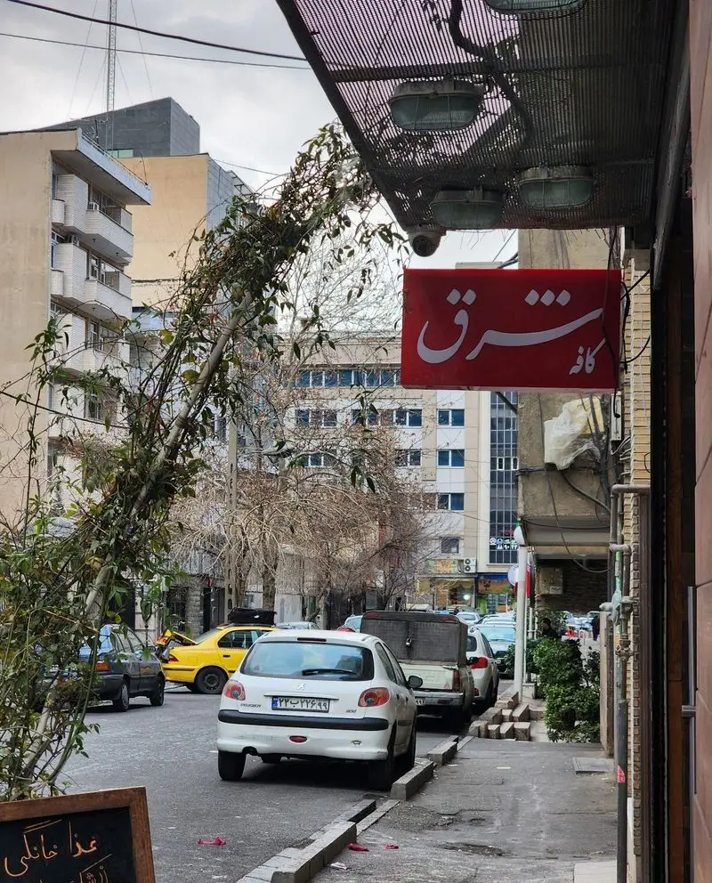 کافه شرق: نمای بیرونی کافه با تابلوی قرمز رنگ در خیابان، همراه با ماشین‌های پارک شده و فضای سبز شهری در تهران.