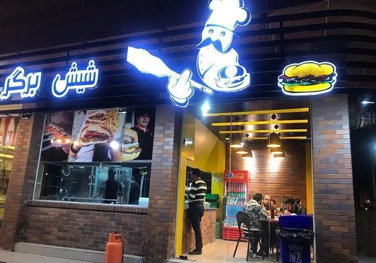 تصویری از نمای شب فست فود شیش برگر: تابلوی نئون با نام شیش برگر، لوگو سرآشپز، و عکس های جذاب برگر و مشتریان.