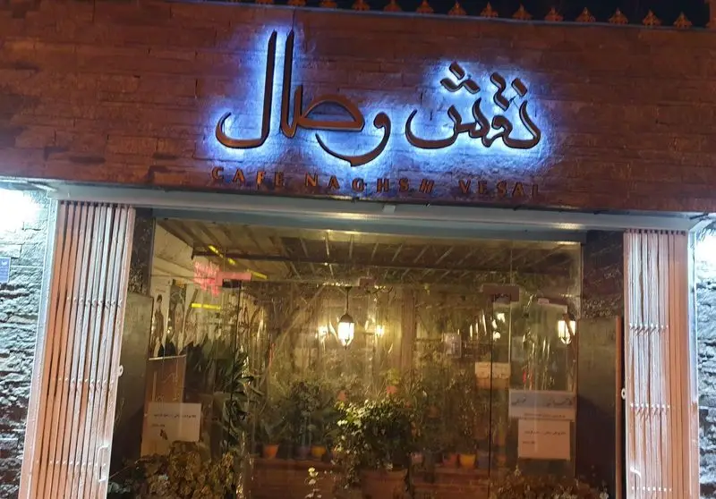 عکس شب از نمای بیرونی کافه نقش وصال با تابلوی نورانی جذاب "کافه نقش وصال" به فارسی و انگلیسی روی دیوار آجری. ورودی شیشه‌ای کافه با فضای داخلی سرسبز.
