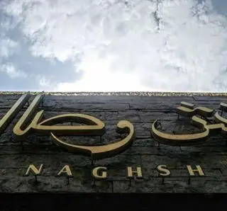 تابلوی کافه نقش وصال: حروف برجسته طلایی "نقش وصال" به فارسی و "NAGHSH" به انگلیسی، روی دیوار سنگی زیر آسمان ابری.