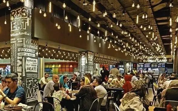 تصویری شلوغ از فضای داخلی رستوران فست فود برگرلند با طراحی مدرن و چراغ‌های آویز زیاد و مشتریانی در حال صرف غذا.