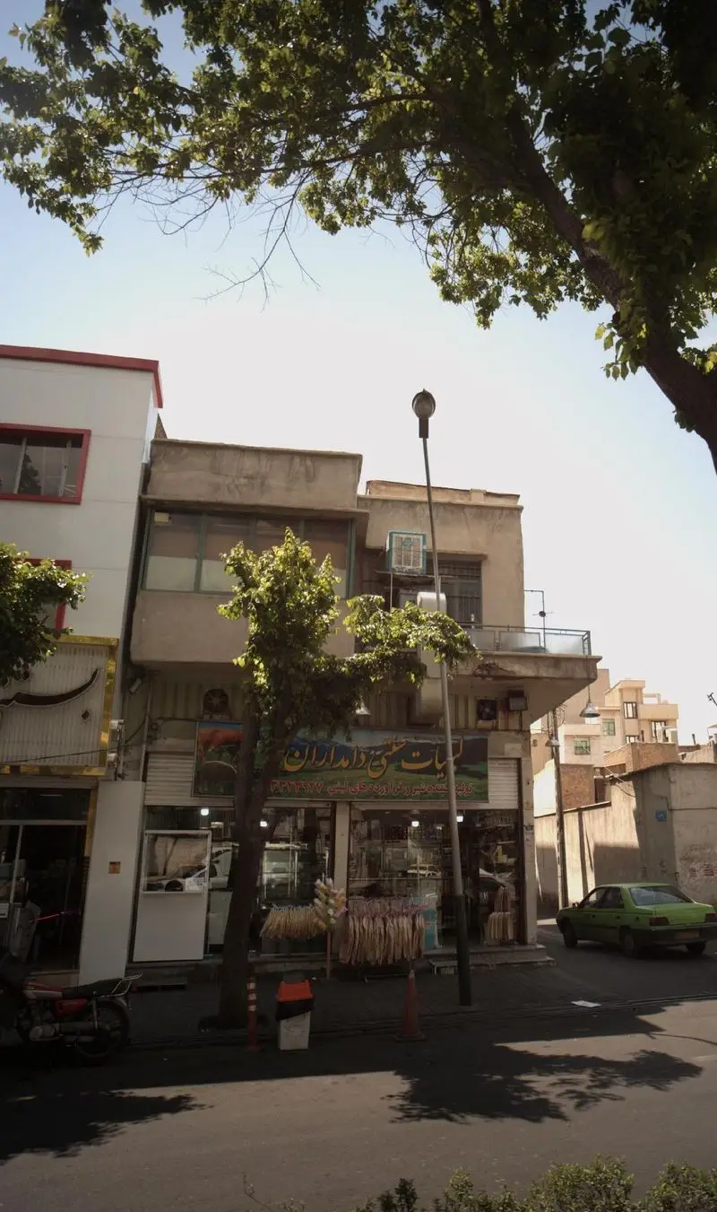 ورودی فروشگاه لبنیات سنتی دامداران در یک روز آفتابی، نمای نزدیک از محصولات لبنی و یک ماشین سبز در حال عبور از خیابان.