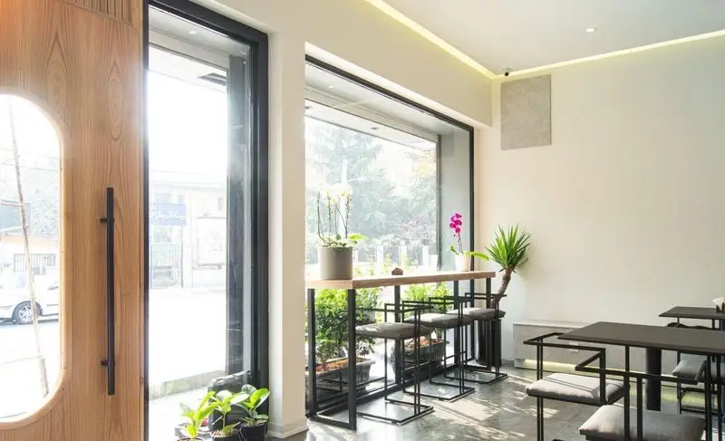 نمای داخلی کافه بردوتی، کافی شاپ مدرن با دکوراسیون دنج، نور طبیعی فراوان از پنجره‌های بزرگ، میز بار چوبی، صندلی‌های شیک و گیاهان سبز.