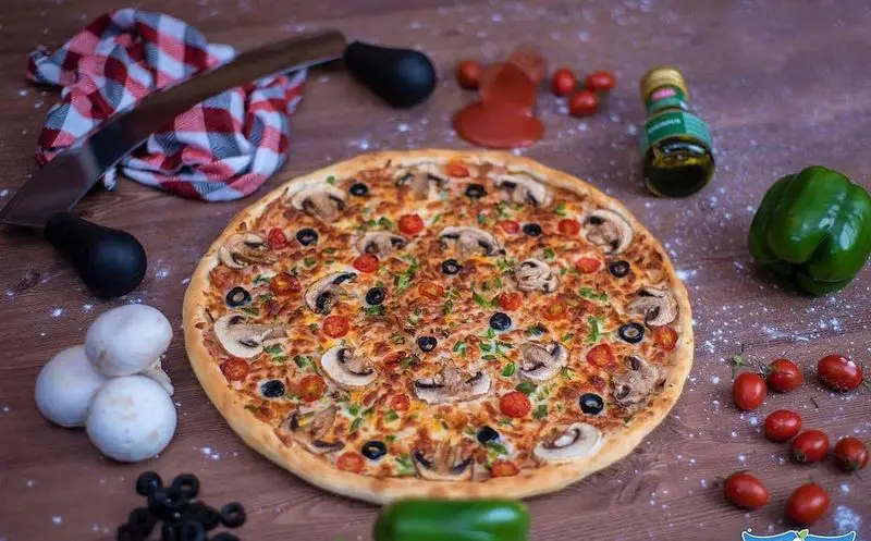 پیتزای خوشمزه قارچ، گوجه گیلاسی و زیتون سیاه از سیب. آماده سازی با مواد اولیه تازه و نمایش جذاب روی میز چوبی رستوران.