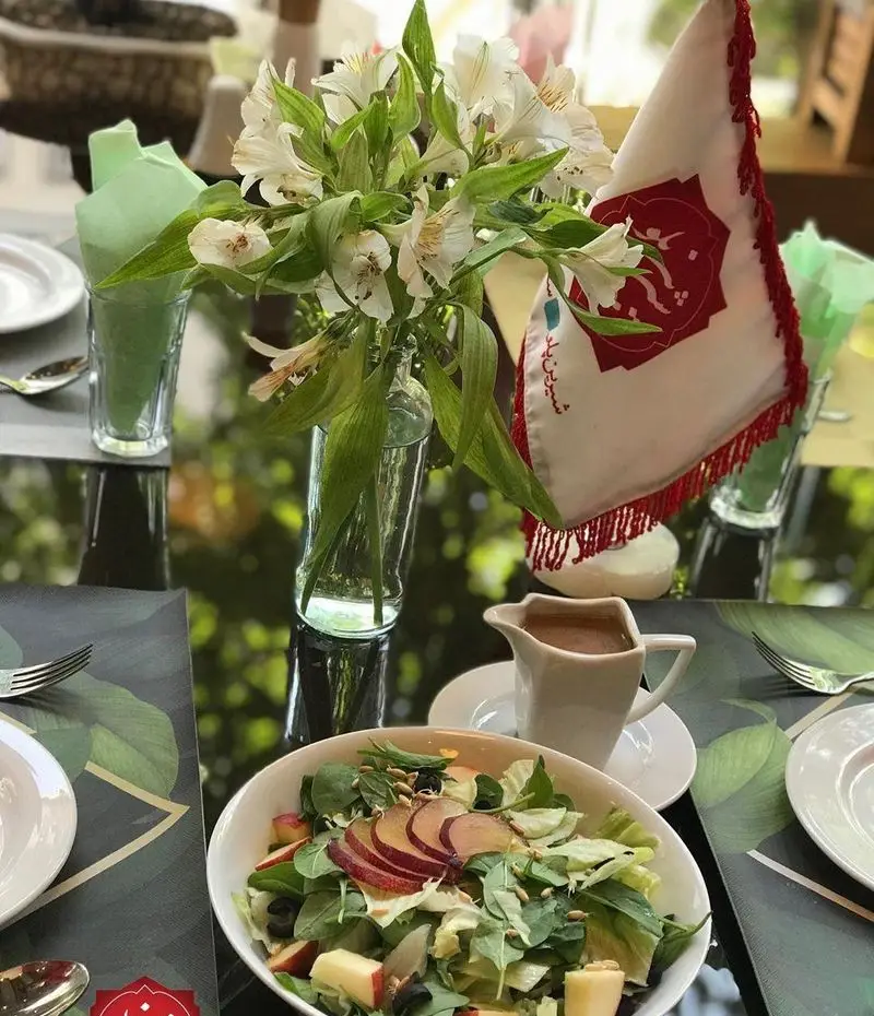 تصویری از میز غذا در رستوران شیرین پلو، شامل سالاد میوه و سبزیجات تازه، سس، گلدان گل‌های سفید زیبا و پرچم برند. فضای دلنشین برای یک وعده سالم.