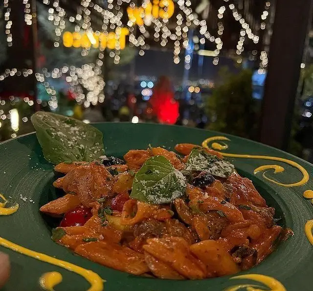 تصویری جذاب از پاستای خوش‌طعم با سس خاص و ریحان، سرو شده در بشقاب سبز کافه رستوران سیتی تراس با دکوراسیون دلنشین و نمای شب شهر.