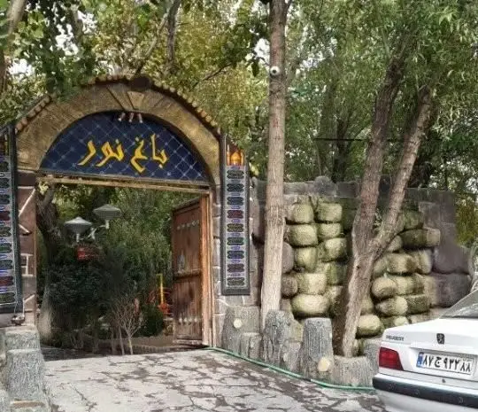 ورودی زیبا و سرسبز کافی شاپ باغ نور با طاق سنگی مزین به نام "باغ نور"، درختان بلند، دیوار صخره‌ای و فضایی دلنشین.