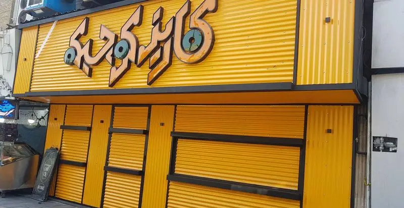 تصویری از نمای خارجی فست فود کاریز کوچیکه، با تابلوی زرد رنگ بزرگ و روشن که نام رستوران را به خطی زیبا نشان می‌دهد. کرکره‌های زرد و قاب‌های مشکی به آن جذابیت بخشیده‌اند.