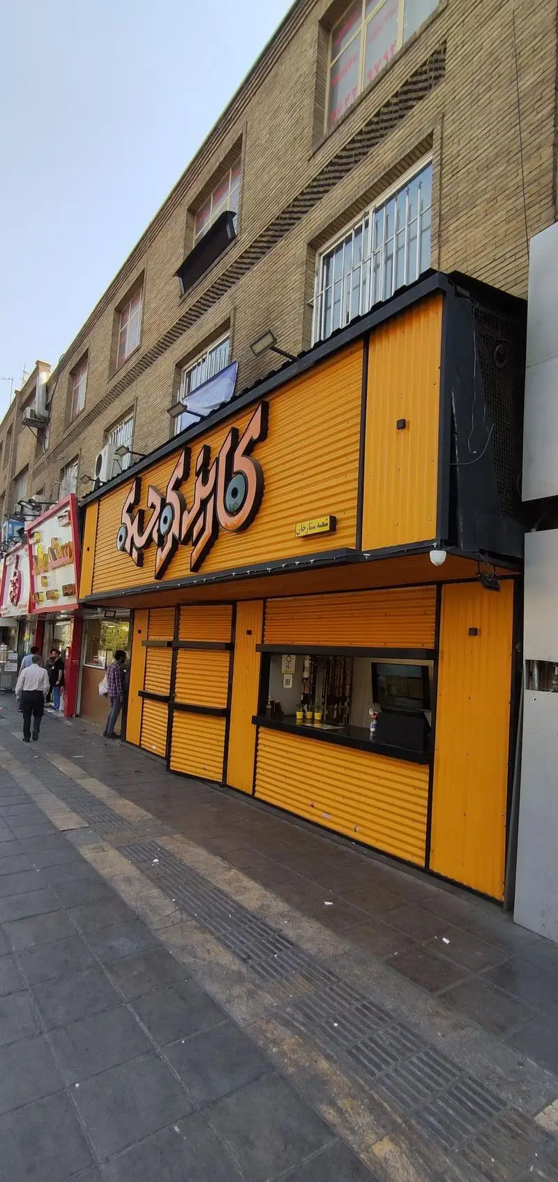 فست فود کاریز کوچیکه با نمای نارنجی روشن، تابلو بزرگ و طراحی مدرن در کنار خیابان، با عابران پیاده و ساختمان‌های آجری قدیمی.