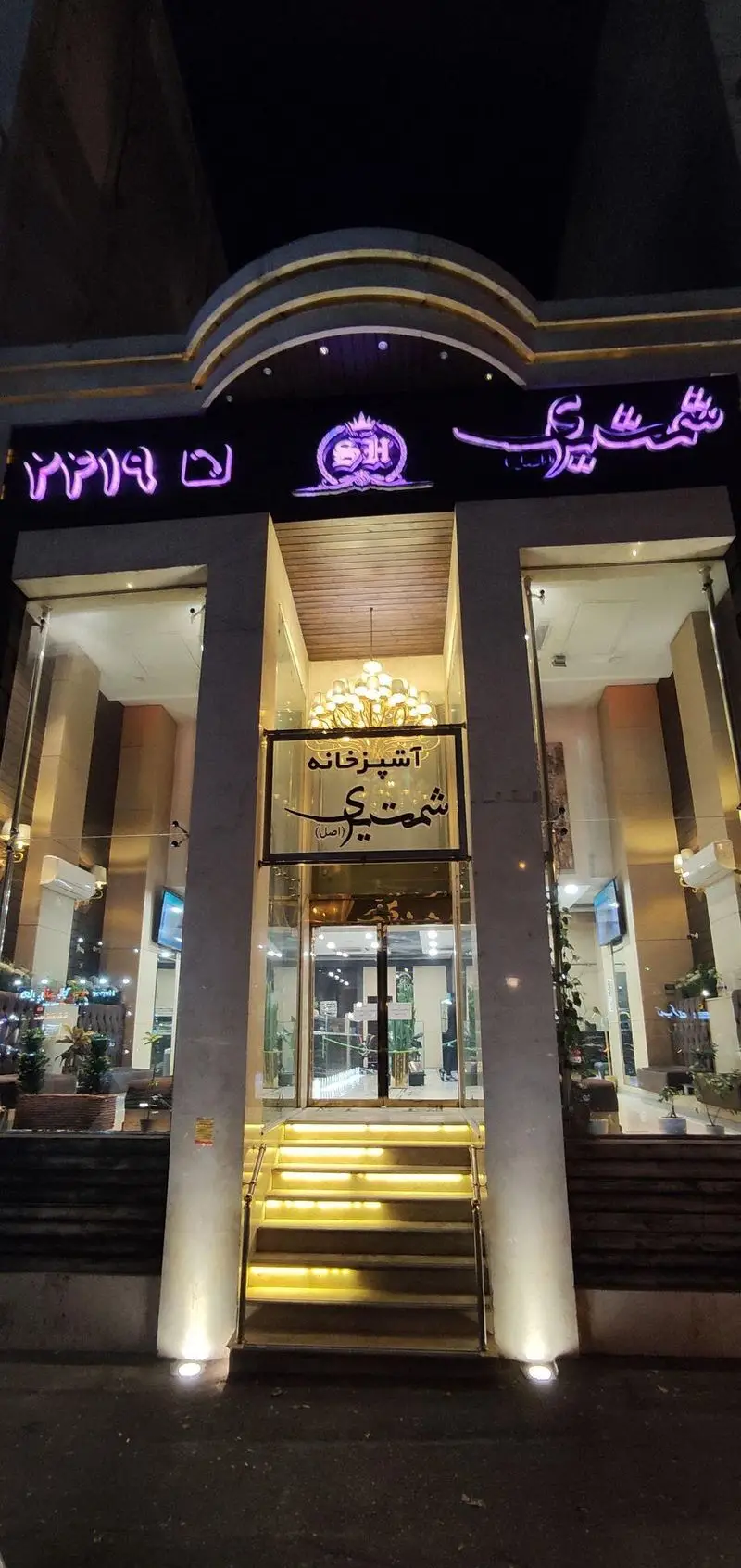 نمای شب و ورودی مجلل آشپزخانه و رستوران شمشیری (اصل) با تابلوهای نئونی درخشان، پله‌های نورانی و سردر شیک، آماده پذیرایی در فضایی دلنشین.