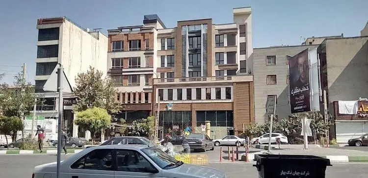 نمایی از خیابان شلوغ شهری با ساختمان‌های چند طبقه، خودروها و عابران پیاده در نزدیکی رستوران شمشیری. بیلبورد شهاب مظفری نیز دیده می‌شود.