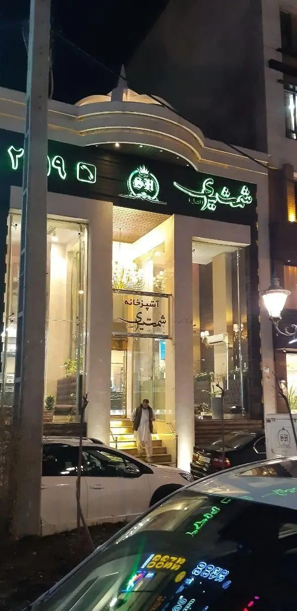 نمای شبانه از ورودی و نمای خارجی رستوران شمشیری با نورپردازی نئون سبز، تابلو آشپزخانه شمشیری و لوگوی SH، محیطی مدرن و شیک.