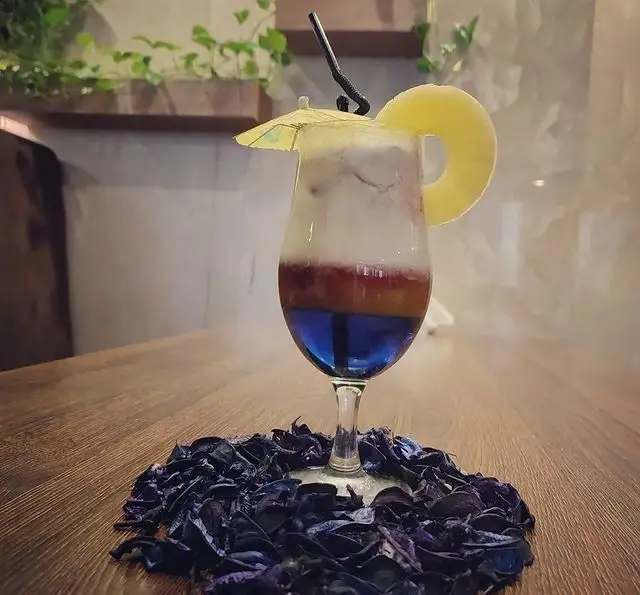 نوشیدنی کوکتل لایه‌ای و رنگارنگ سفره خانه خیام با تزئین آناناس، چتر و گلبرگ‌های خشک، خنک و دلنشین برای روزهای گرم.