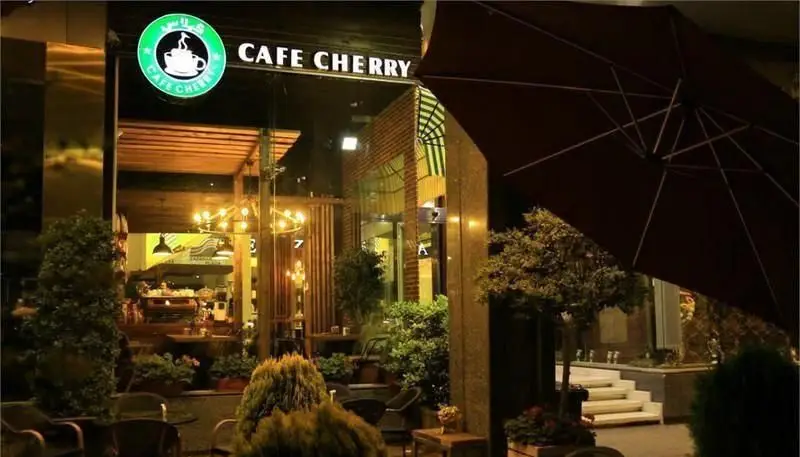 نمای بیرونی کافه چری در شب با نورپردازی گرم و محیطی دنج، لوگوی سبز روشن و میز و صندلی‌های فضای باز جذاب.
